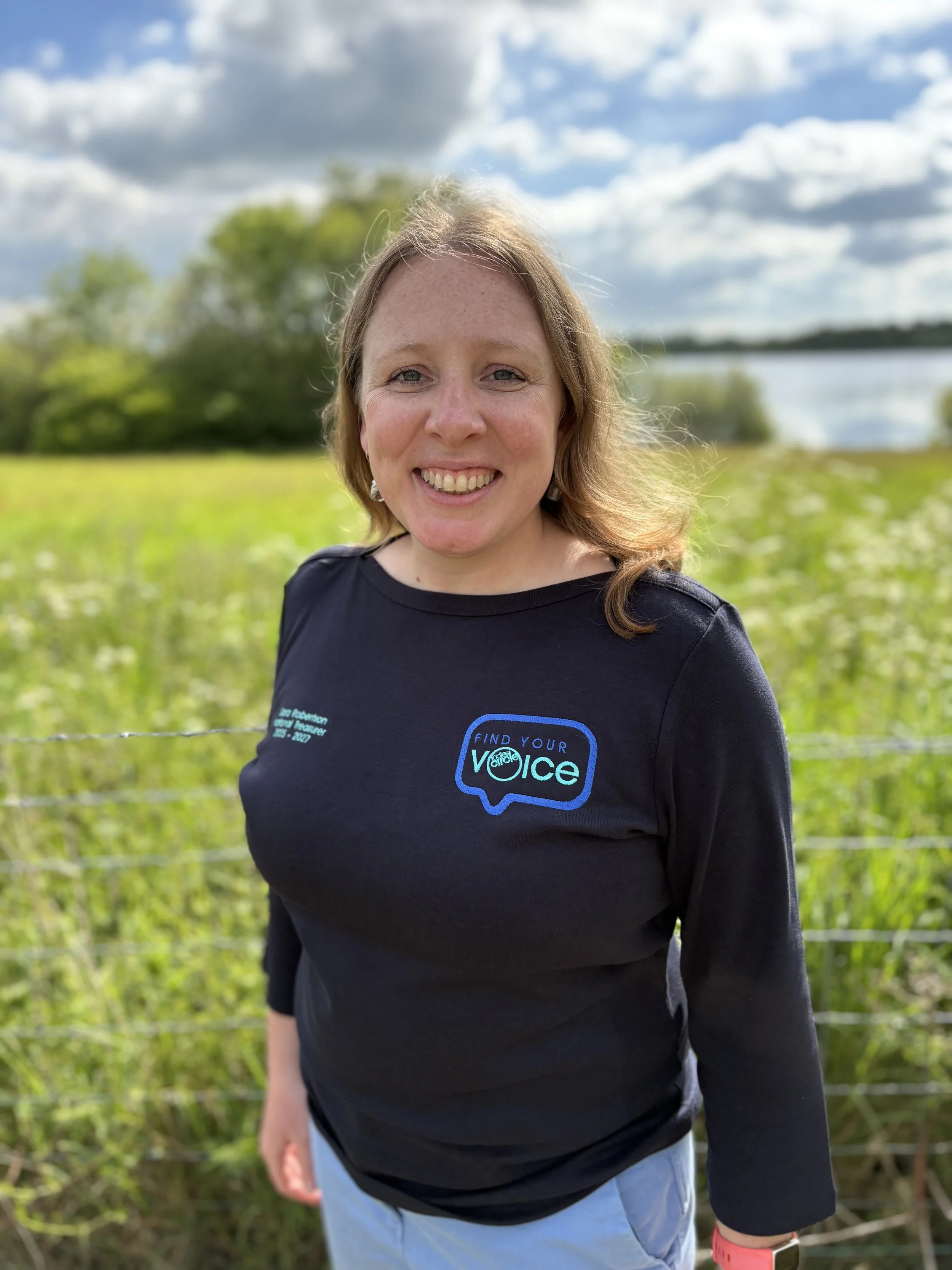 A woman with reddish-brown hair and light skin smiling outdoors, wearing a black shirt with a graphic that says "Find Your Voice" and a speech bubble icon, standing in front of green vegetation near a river under a partly cloudy sky.