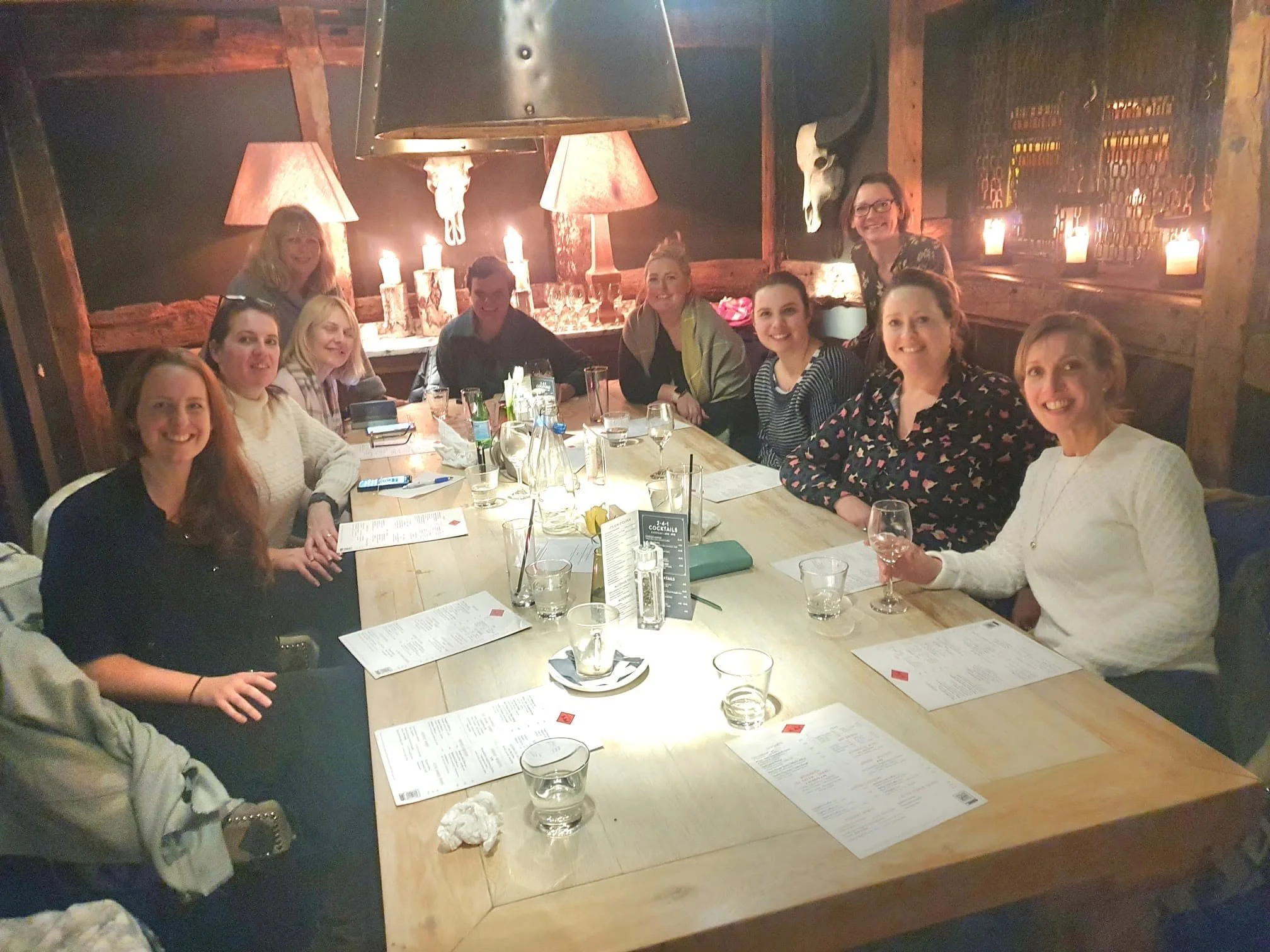 A group of ten women sitting around a wooden table in a cozy, rustic restaurant with dim lighting, candles, and decorative elements like lamps and a skull on the wall.