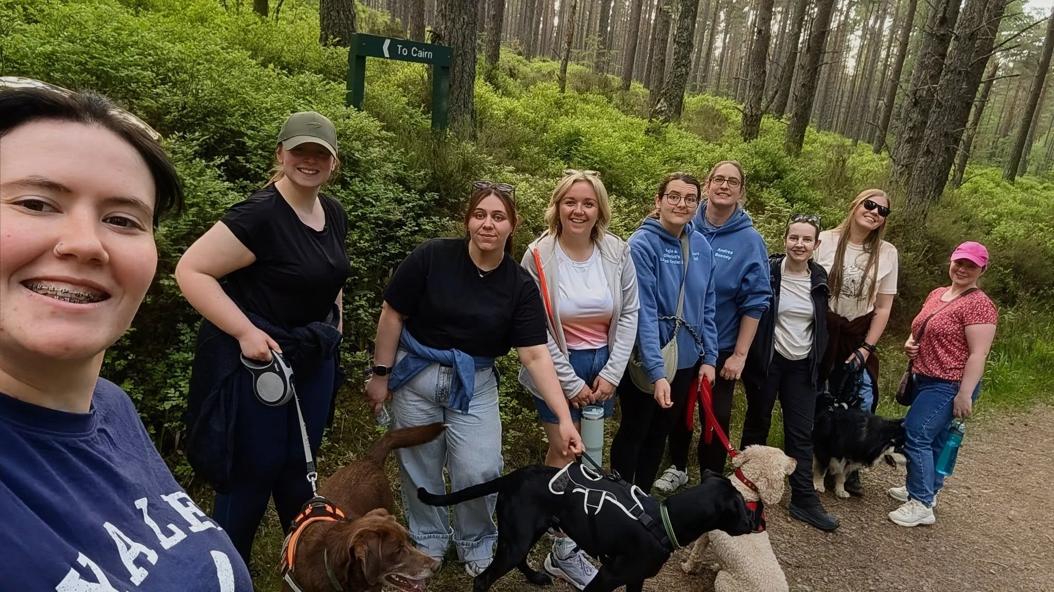 A group of women and dogs on a forest trail during daytime, smiling and posing for a photo amidst trees and lush greenery.