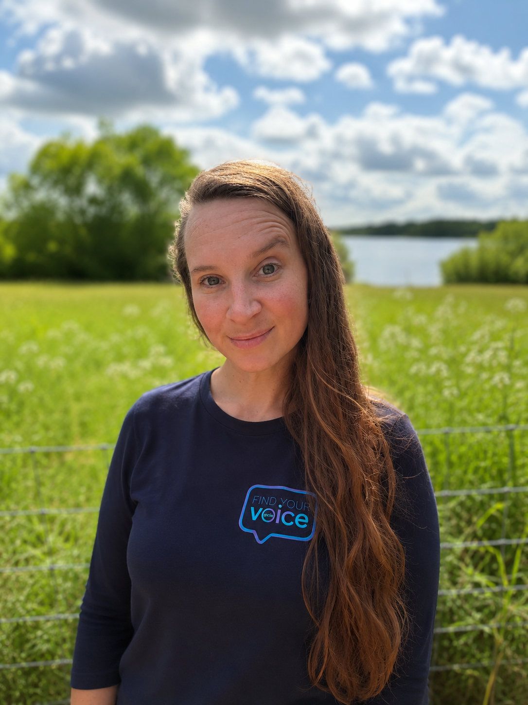 A woman with long brown hair smiling outdoors in a field, wearing a navy blue shirt with a speech bubble logo that says 'Find Your Voice', with a lake, green trees, and a partly cloudy sky in the background.