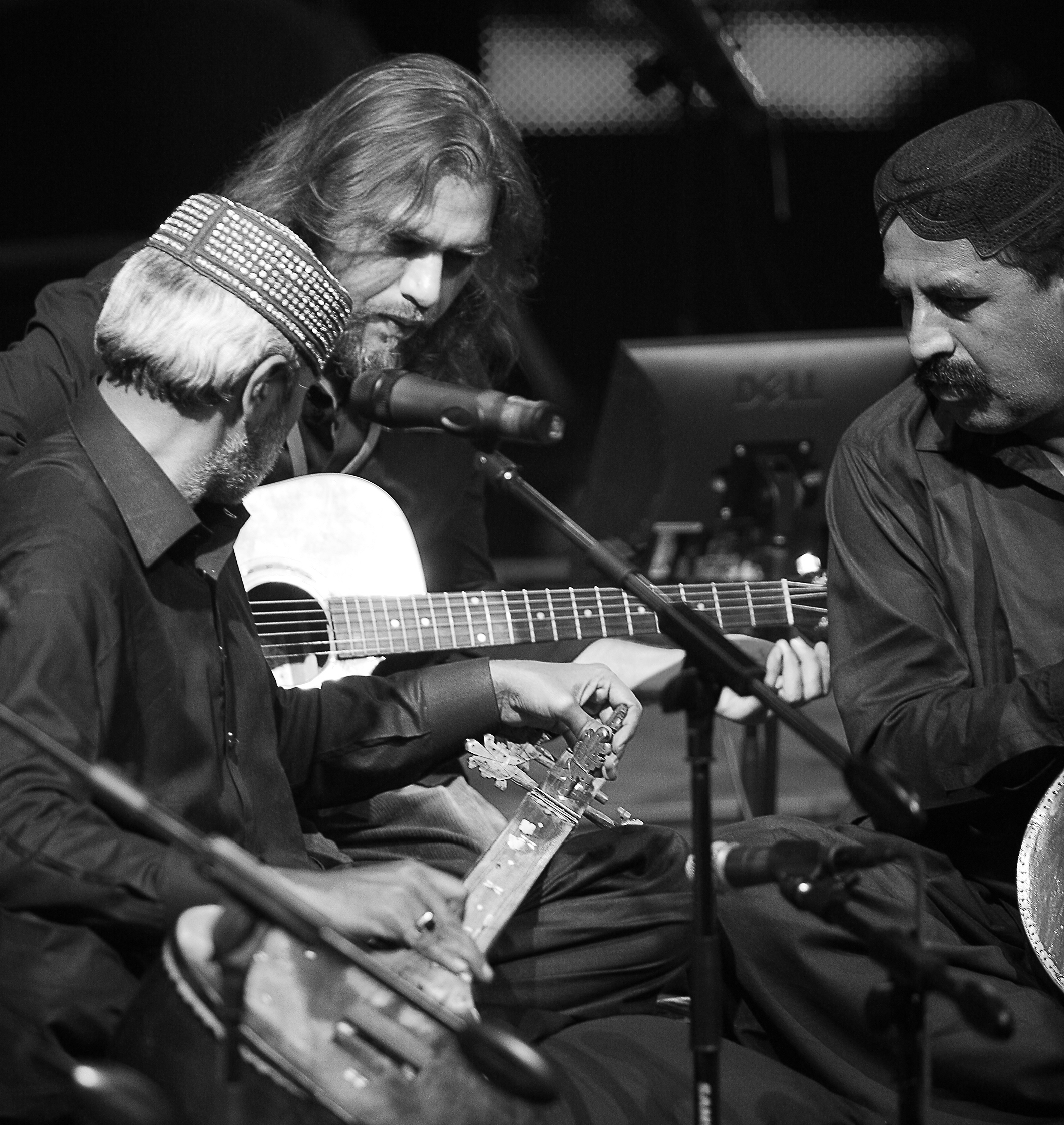 Two men are playing musical instruments in a dark setting. The man on the left, with long hair and a beard, is playing a guitar and singing into a microphone. The man on the right, wearing a cap, is also engaged in the performance.