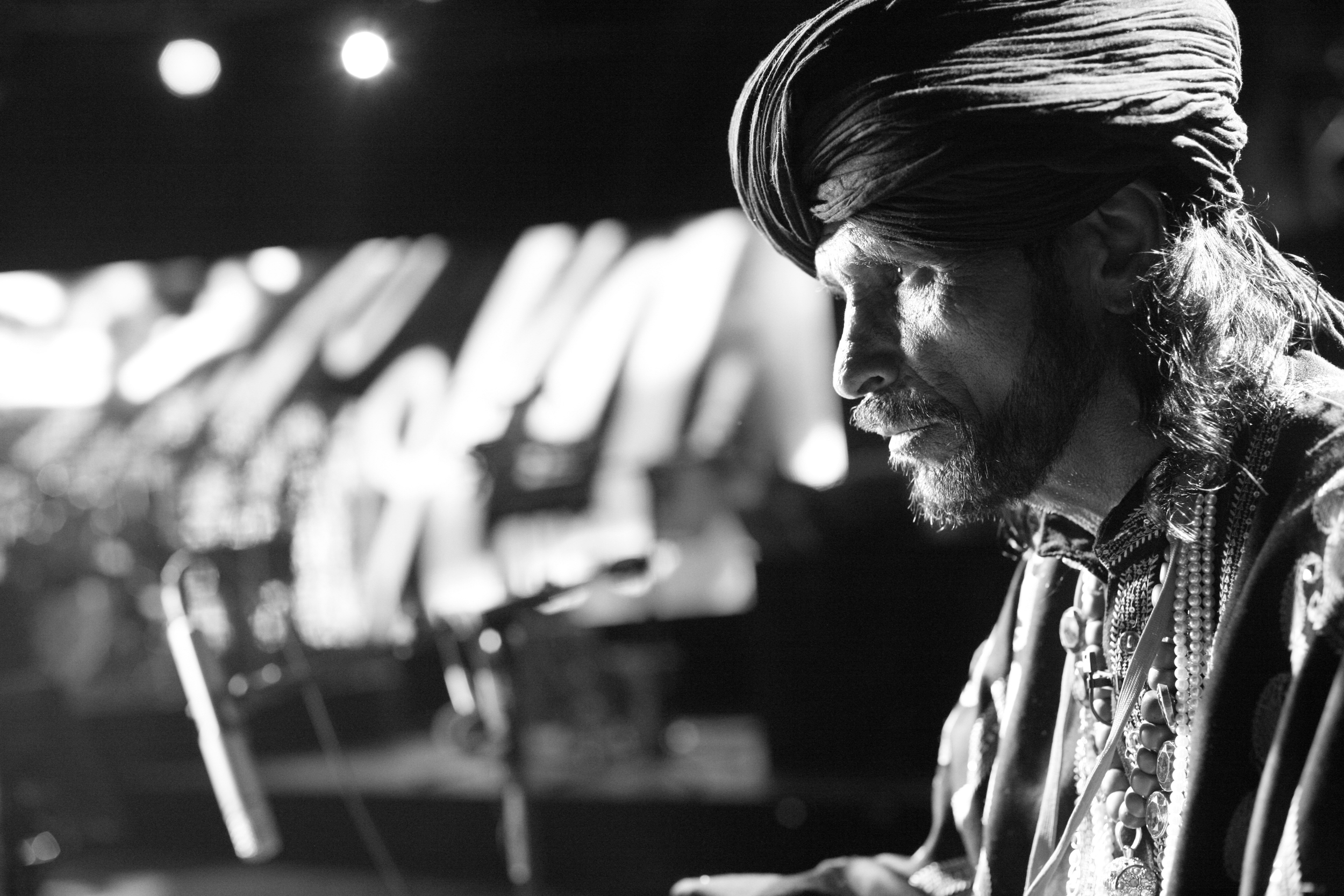 A black and white photo of a man wearing a turban and multiple necklaces, with a focused expression, looking downward. The background is blurred with out-of-focus lights and objects.