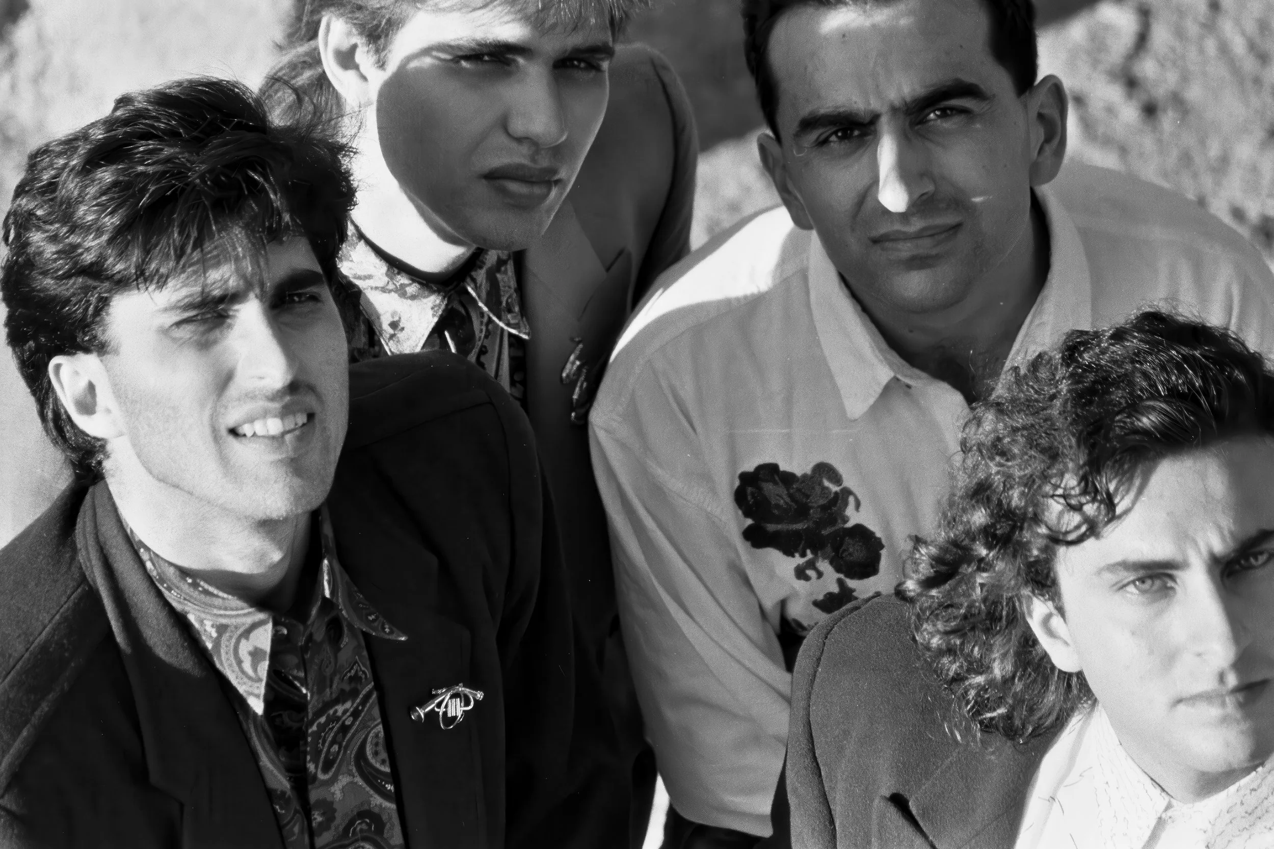 A black and white photo of five young men posing outdoors, with some looking directly at the camera and others slightly away. They are dressed in a variety of casual and semi-formal clothing, with rocky terrain visible in the background.