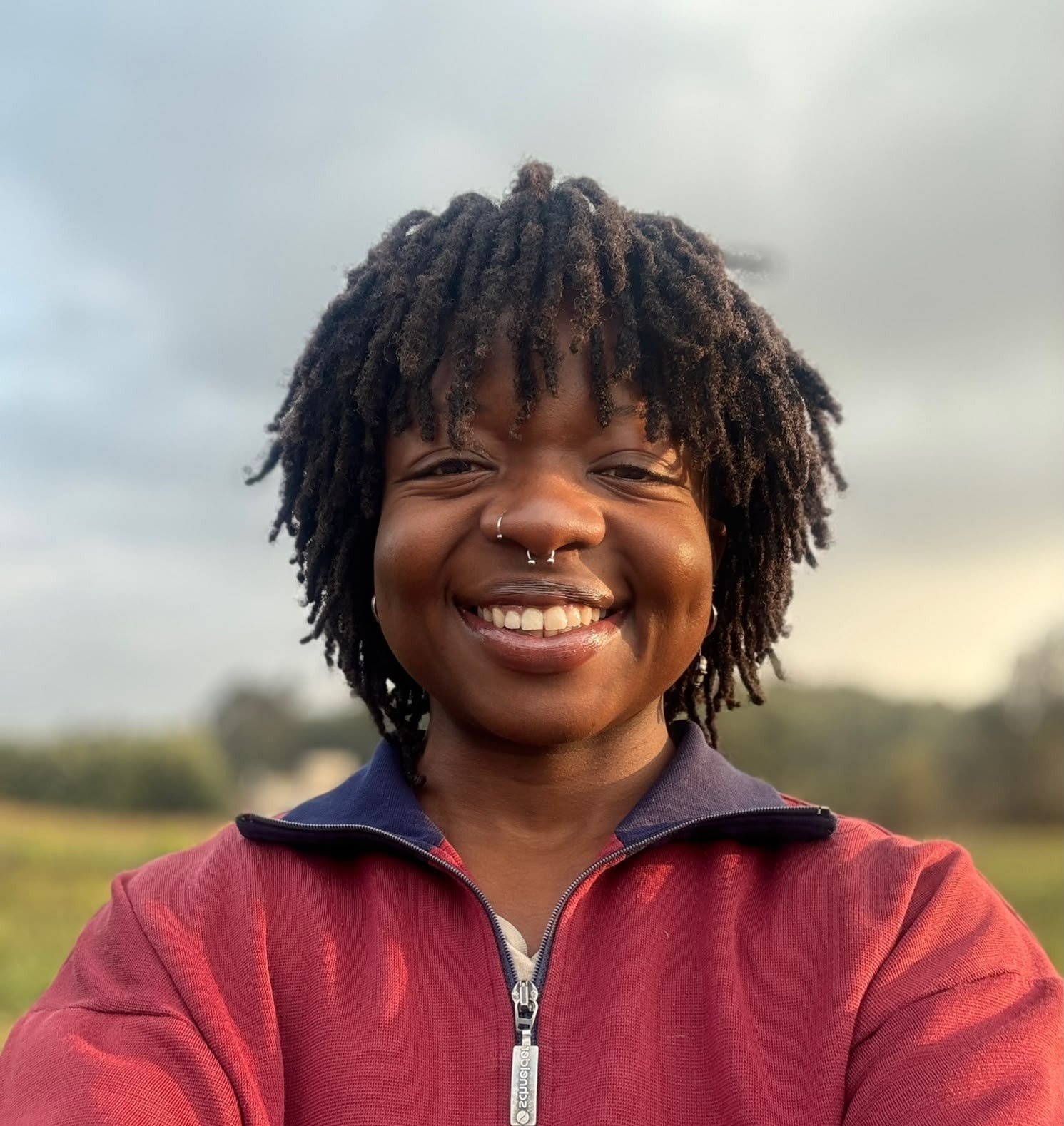 A woman with short, curly dreadlocks and nose piercings taking a selfie outdoors under cloudy skies, wearing a red zip-up jacket and smiling.