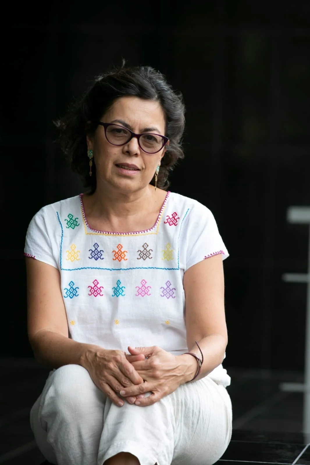 A woman with dark wavy hair, wearing glasses, a white embroidered blouse with colorful patterns, and white pants, sitting against a black background.