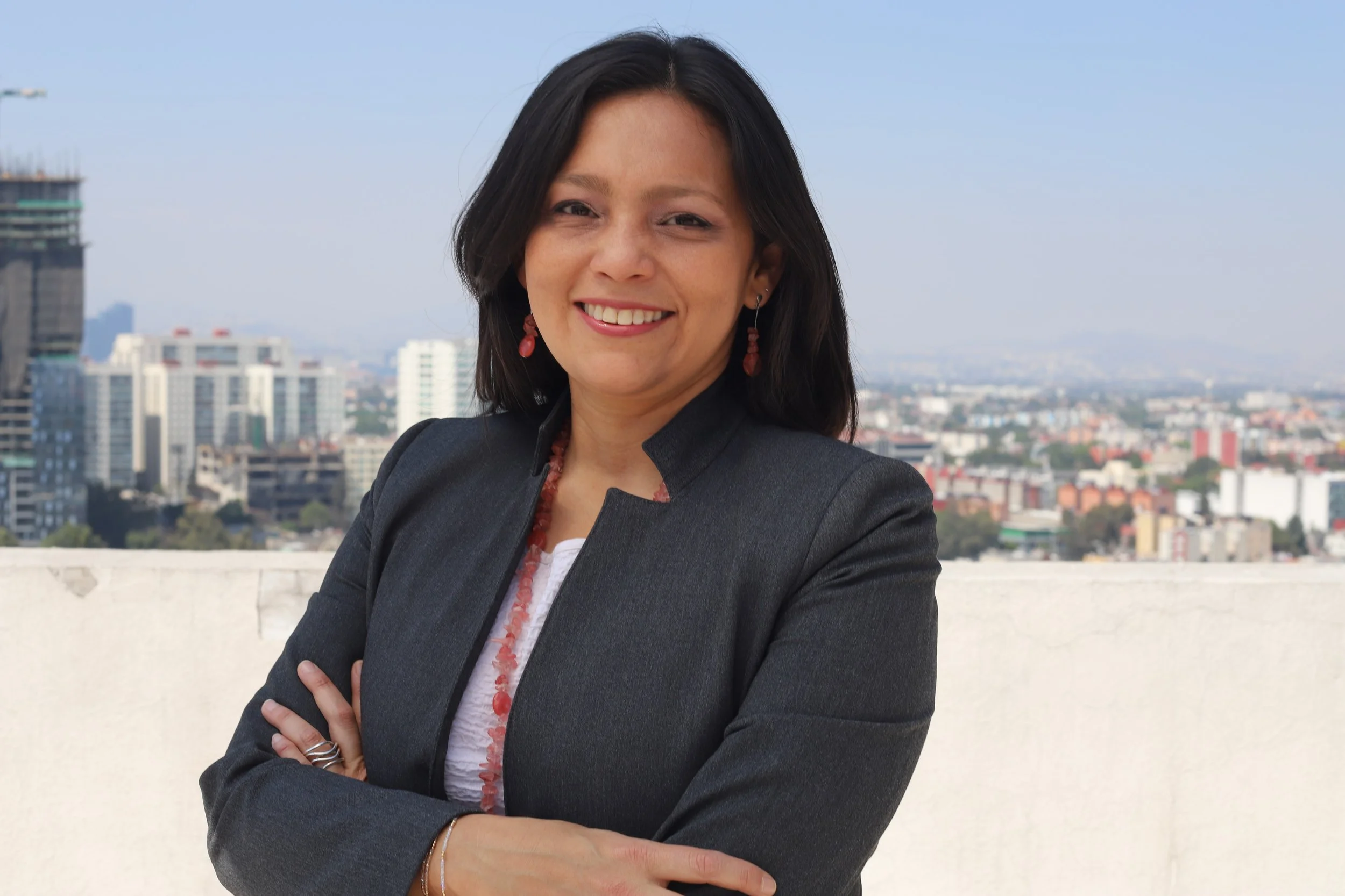 Portrait of a smiling woman with dark shoulder-length hair, wearing earrings, a dark blazer, and a necklace, standing outdoors with a city skyline in the background.