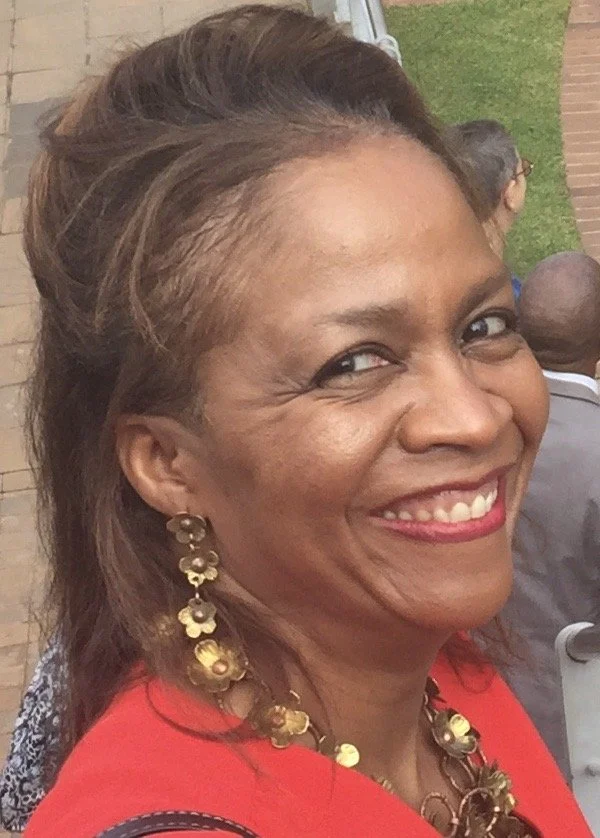Close-up of a smiling woman with short, styled hair, wearing gold jewelry and a red top, outdoors at a social gathering.