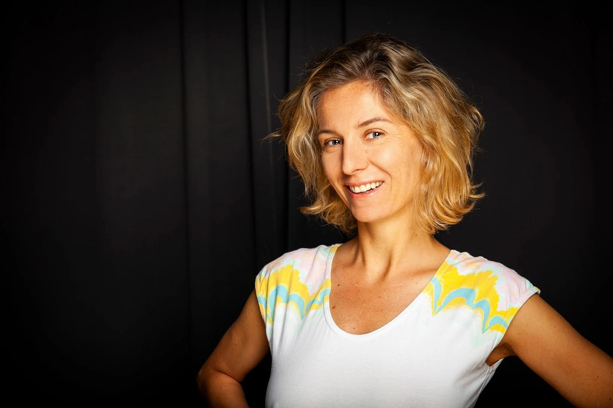 A woman with short blond wavy hair smiling and wearing a white shirt with colorful yellow, blue, and pink patterns.