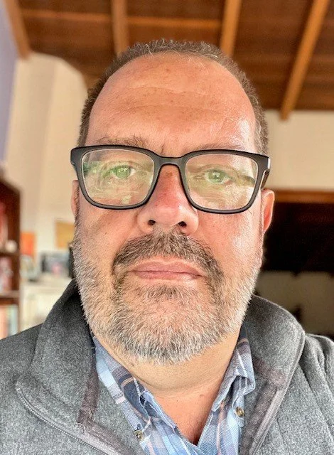 Close-up of a middle-aged man with glasses, a beard, and a gray hoodie inside a room with wooden ceiling beams.