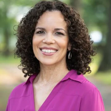A woman with curly hair wearing a purple blouse outdoors, smiling at the camera.