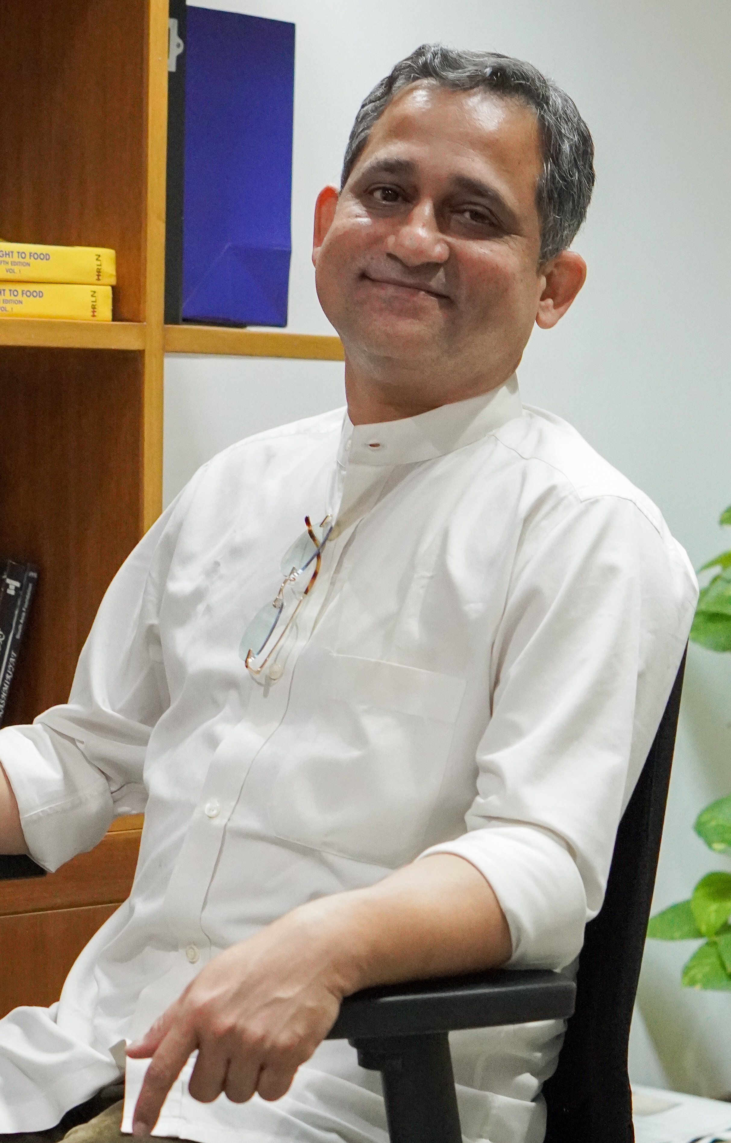A man smiling, wearing a white shirt, sitting in a black office chair, with a bookshelf and colorful boxes in the background.