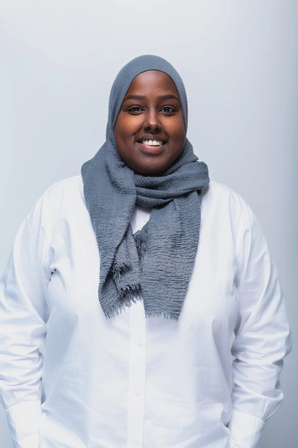 A woman wearing a gray headscarf and a white shirt, smiling at the camera against a plain background.