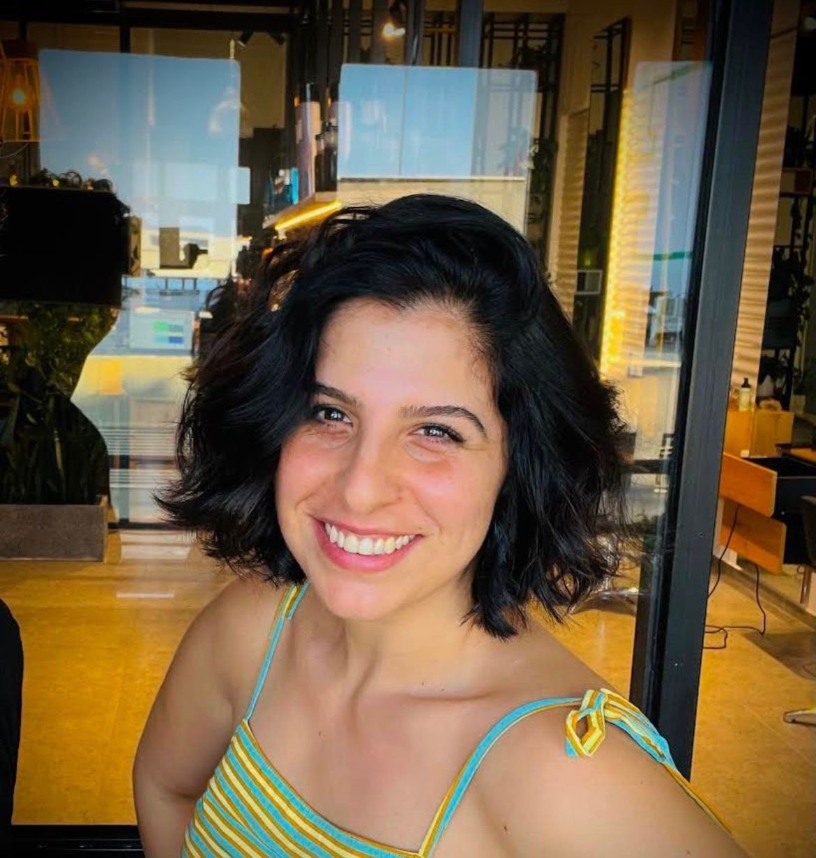A woman with shoulder-length dark curly hair smiling, wearing a colorful striped summer dress with shoulder ties, in a modern indoor space with large glass windows and warm lighting.