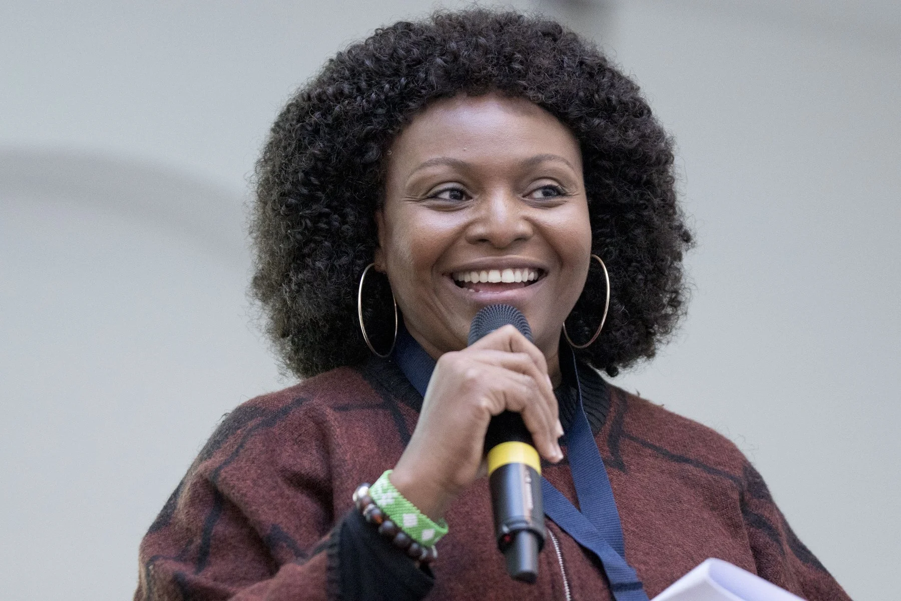 A woman with curly black hair, wearing hoop earrings and a burgundy checkered sweater, speaking into a microphone at an indoor event.