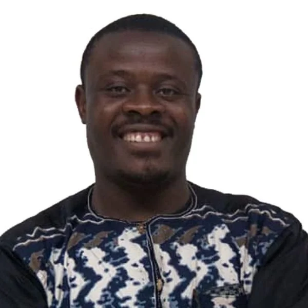 A man with dark skin and short black hair, smiling, wearing a patterned shirt and a black jacket, against a white background.