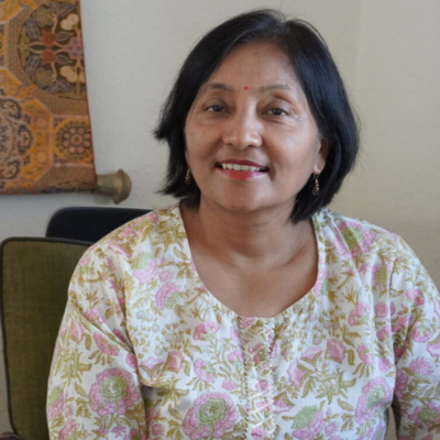 A woman with short black hair, wearing a floral patterned blouse, smiling while sitting indoors.
