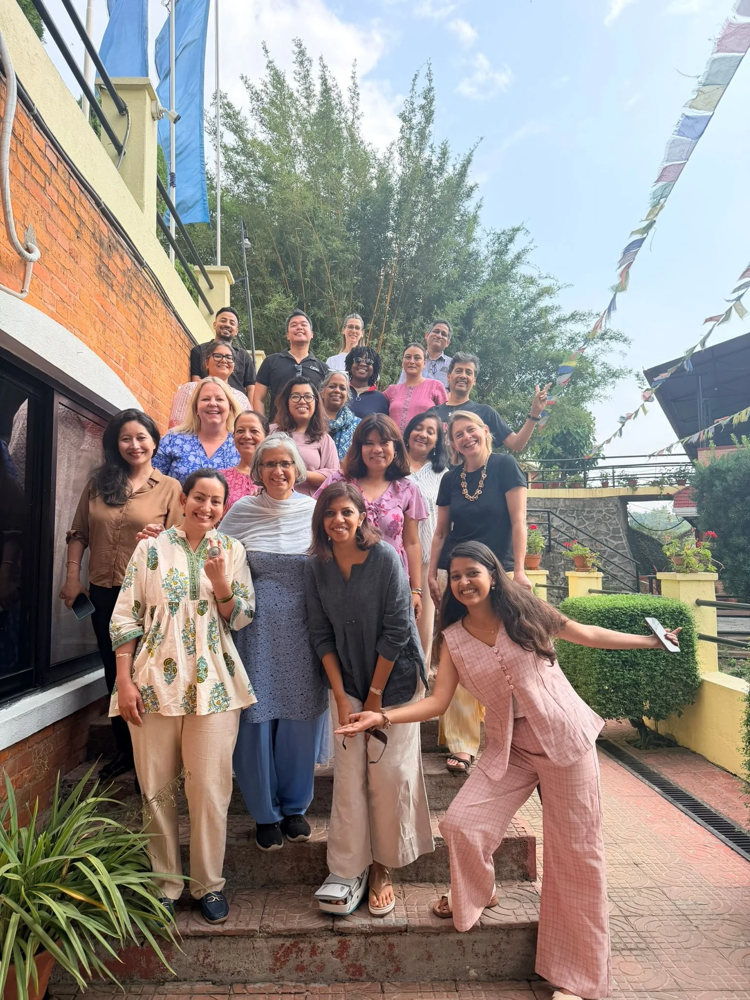 A group of people smiling and posing for a photo outdoors, with some standing on stairs and others on the ground, in a courtyard with greenery and residential buildings.