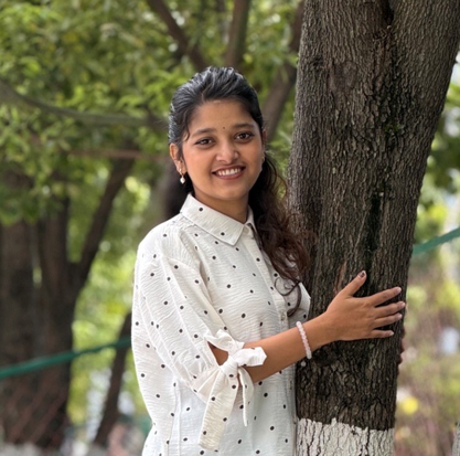 Smiling young woman in a white polka-dot shirt hugging a tree outdoors with green foliage in the background.