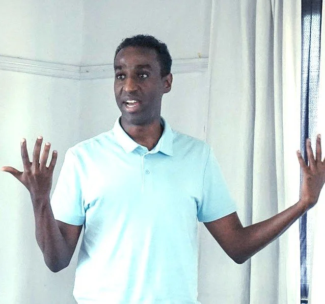 A man in a light blue polo shirt standing indoors with his arms raised and hands open, speaking or gesturing.