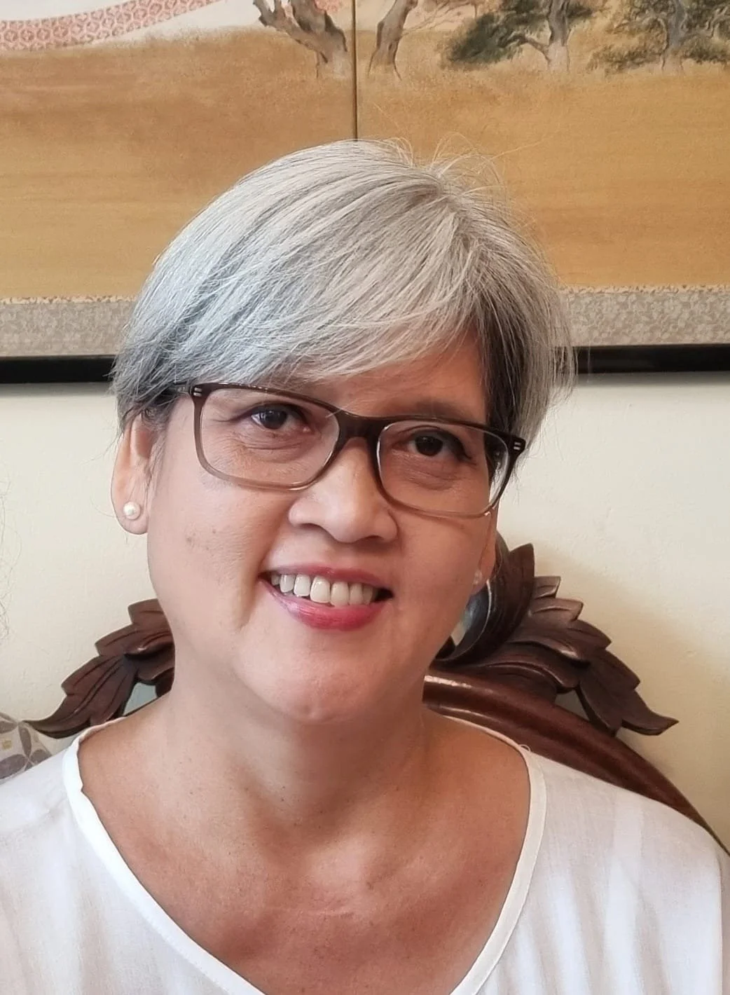 A smiling middle-aged woman with short gray hair, wearing glasses, pearl earrings, and a white top, sitting in front of a wooden chair with a carved backrest, with a painting of trees hanging on the wall behind her.