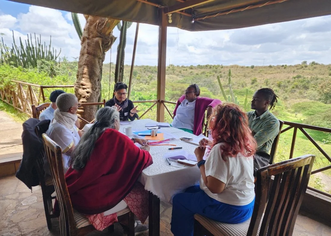 Group of eight people sitting around a table outdoors on a covered deck, engaged in discussion. The background features a large tree, green vegetation, and an open landscape with hills under a cloudy sky.