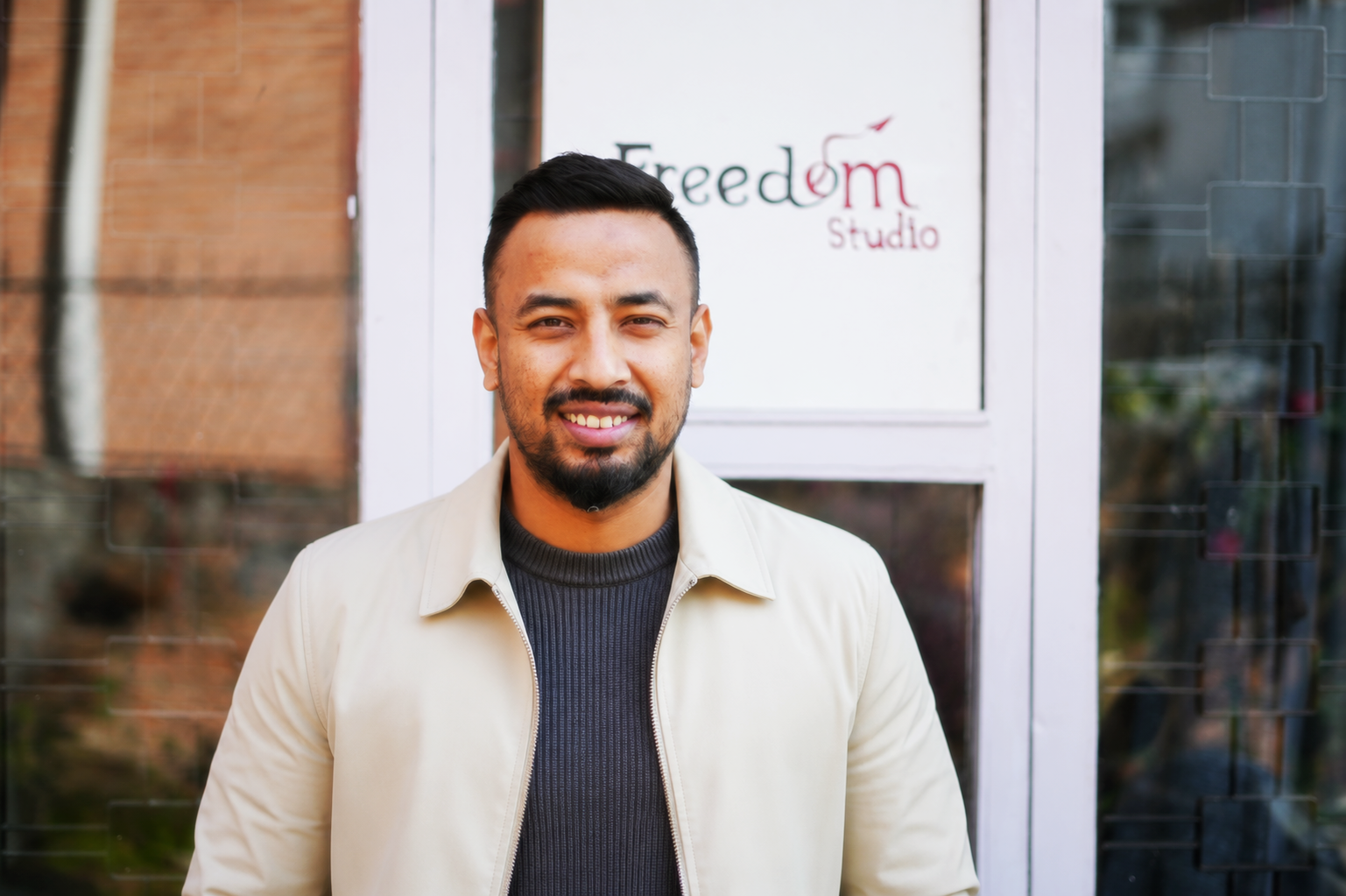 A smiling man with short dark hair and a beard stands in front of a glass door with a sign that says 'Freedom Studio'.