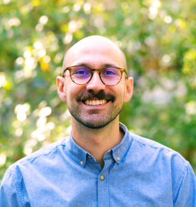 A smiling man with glasses, a mustache, and a beard, wearing a blue button-up shirt, outdoors with greenery in the background.
