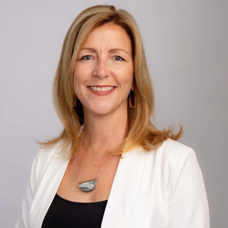 A woman with blonde hair smiling, wearing a black top, white blazer, and a pendant necklace against a plain gray background.