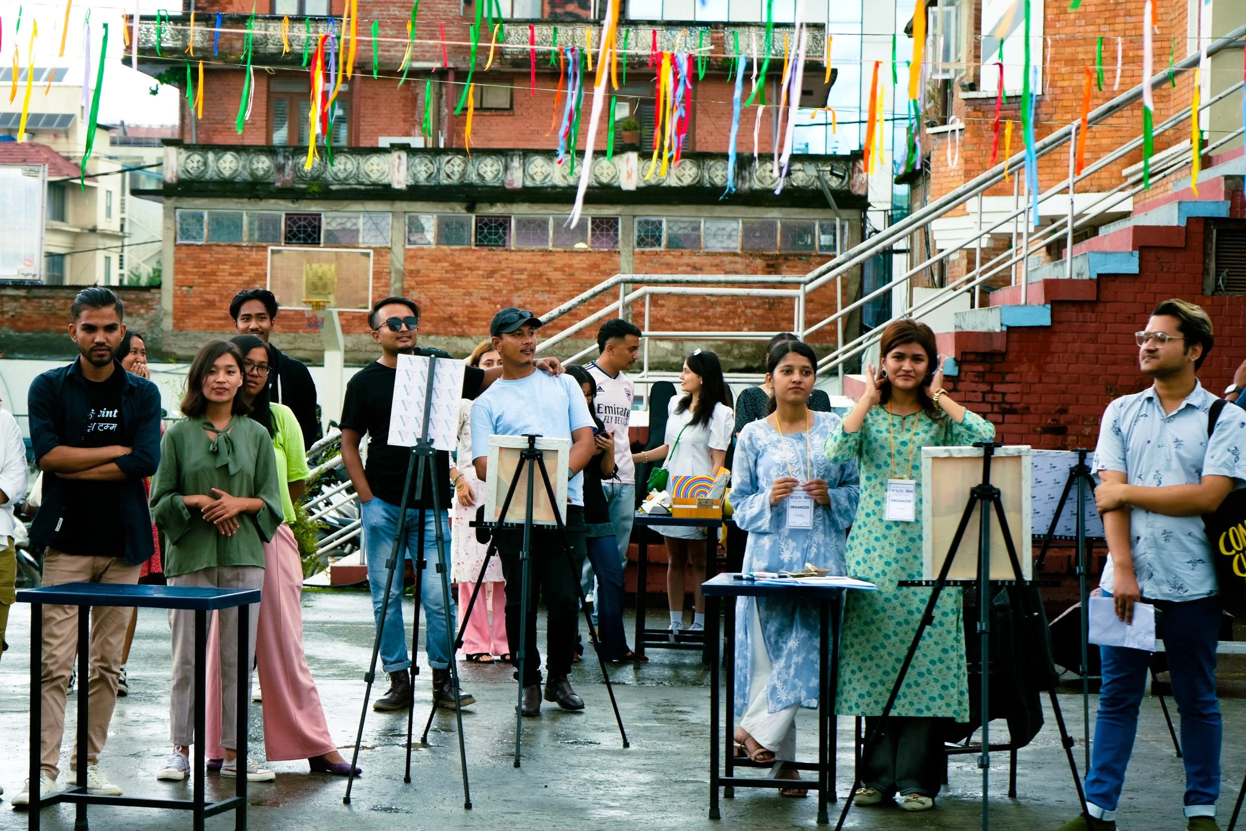 Group of people standing outdoors at a city rooftop or terrace with colorful streamers hanging above. Several tables display artwork, craft items, or informational materials, suggesting an event or gathering.