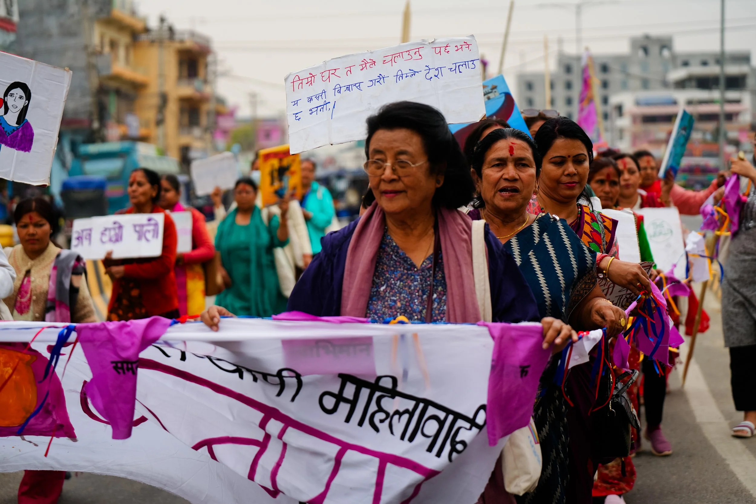 A group of women in traditional attire participating in a protest march on a city street, holding banners and signs.