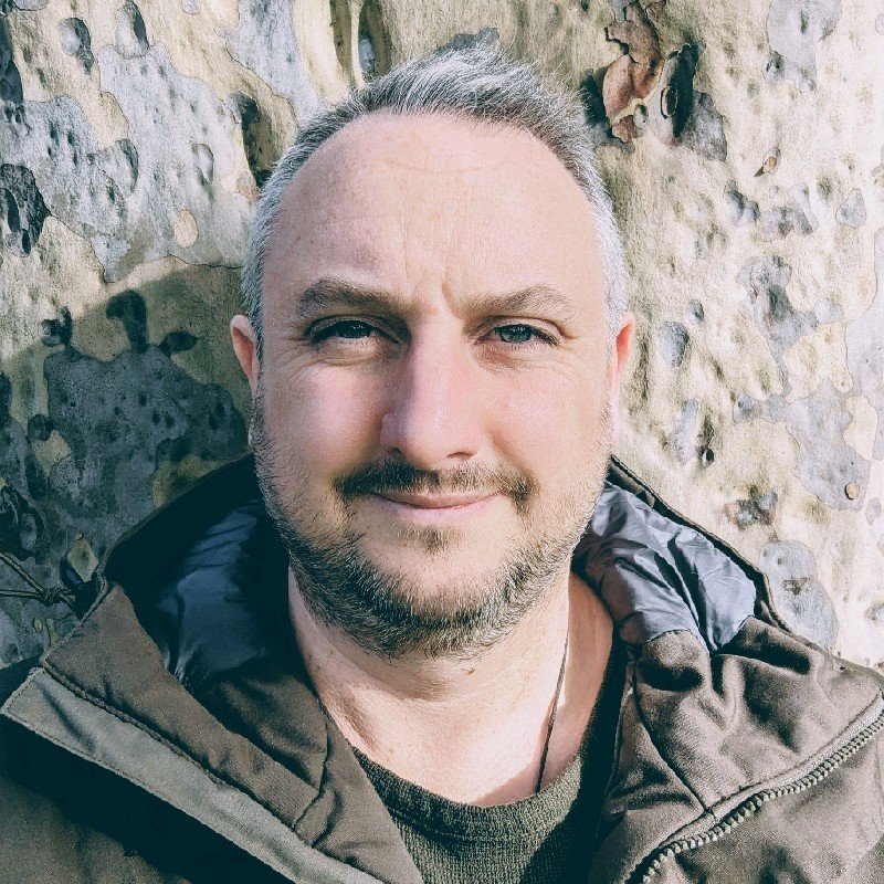 A smiling man with gray hair and a beard taking a selfie outdoors against a stone wall.