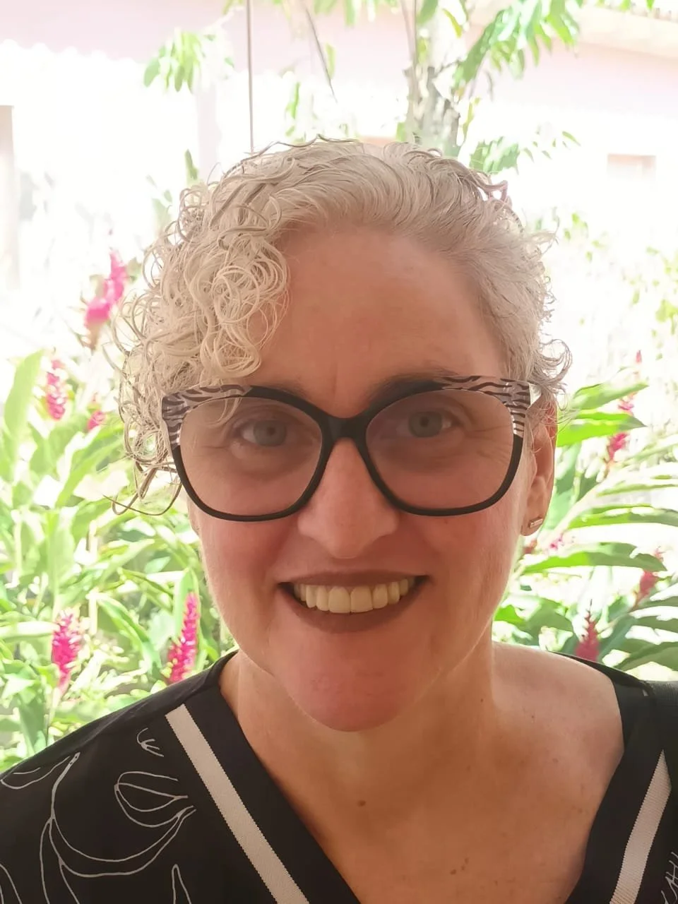 Portrait of a smiling woman with curly blonde hair and black glasses, standing in front of a lush green background with pink flowers.