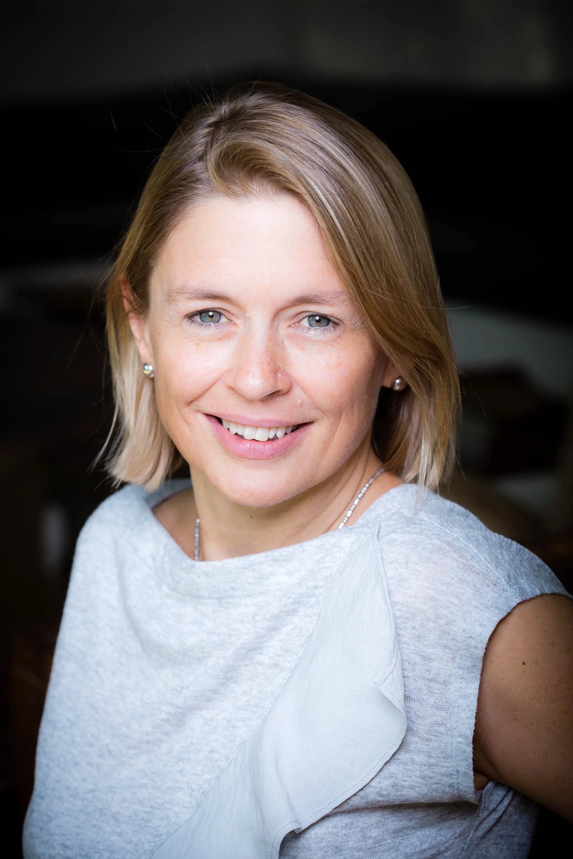 A smiling woman with blonde hair, blue eyes, pearl earrings, and a gray top, posing indoors with a dark background.