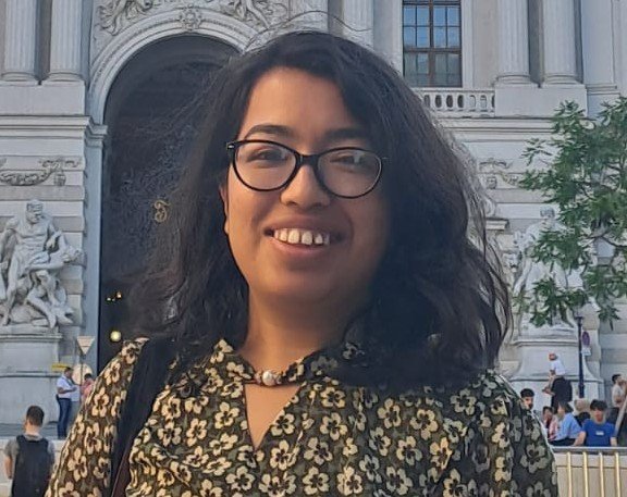 Young woman with glasses and dark wavy hair smiling outdoors in front of a historic building with arched entrance, trees, and people in the background.