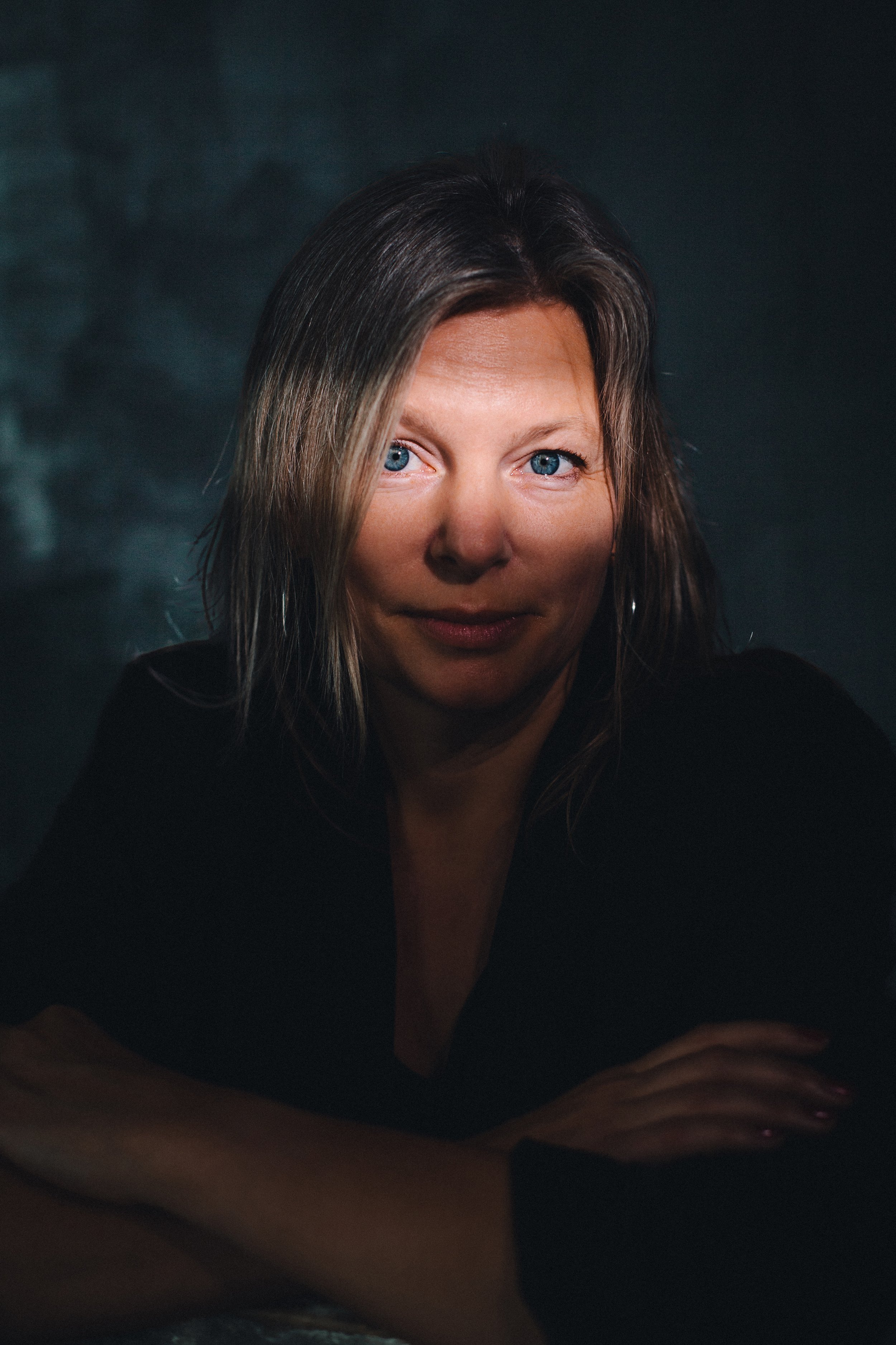Close-up portrait of a woman with blue eyes and dark hair with gray streaks, smiling slightly, wearing a black top, against a dark, blurred background.