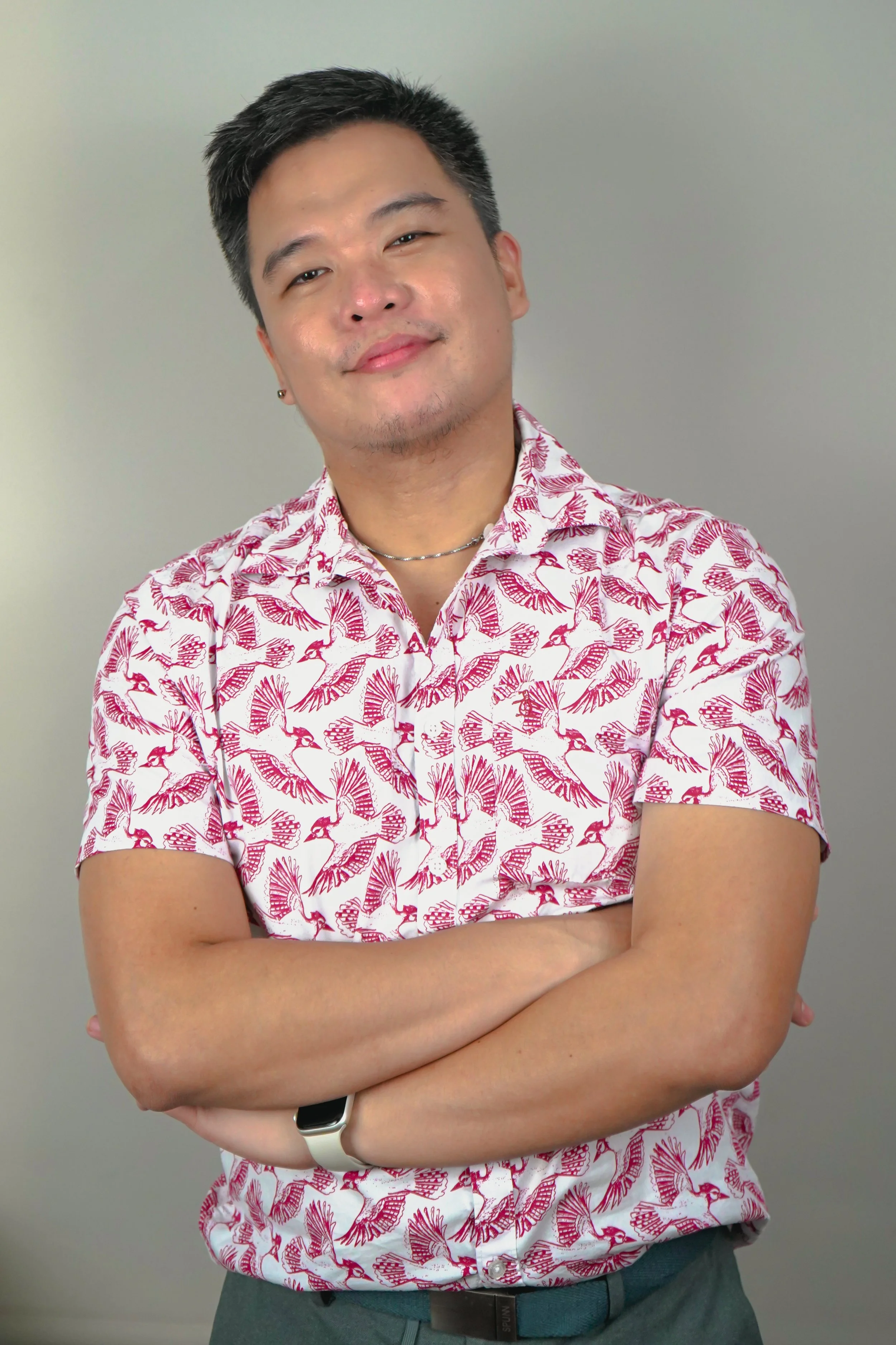 A young man with dark hair, light skin, and a slight smile, standing with his arms crossed in front of a plain gray wall. He is wearing a white shirt with a pink bird print, a silver necklace, a white smartwatch, and a black belt.