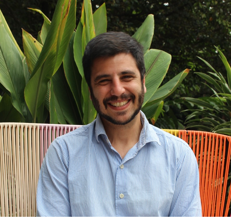 A man with dark hair and beard smiling, sitting outdoors in front of large green leafy plants, wearing a blue and white striped button-up shirt.
