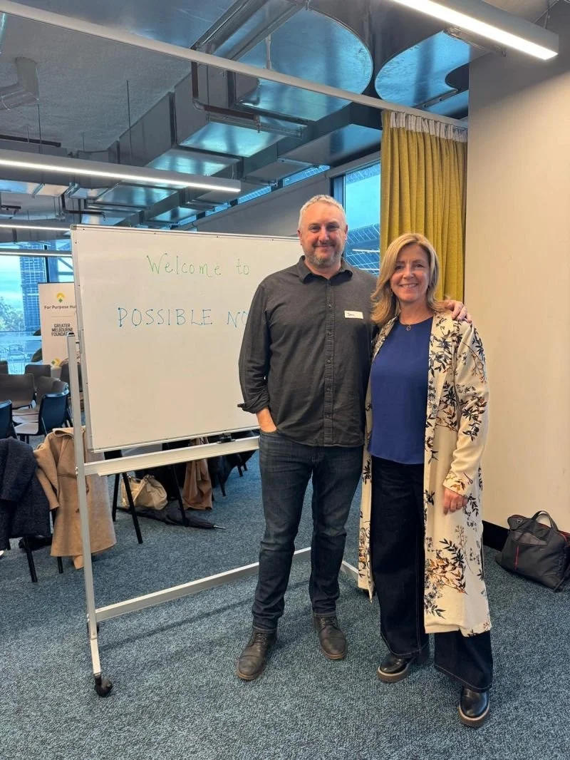 Two people standing next to each other and smiling in an office or conference room, with a whiteboard behind them that reads 'Welcome to POSSIBLE NO' and some chairs and tables in the background.