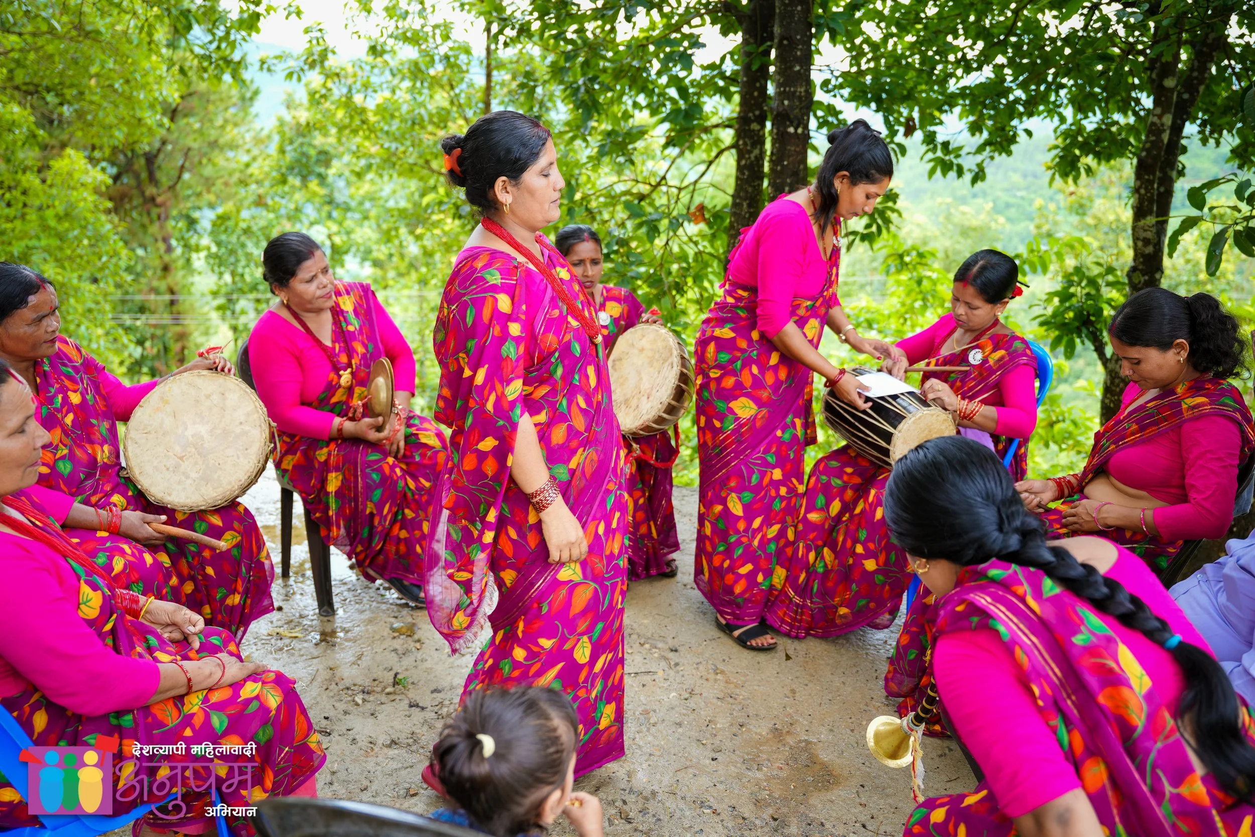 Group of women in colorful traditional clothing, some playing drums and one woman in the center standing with her eyes closed, surrounded by green trees, participating in a cultural outdoor event.