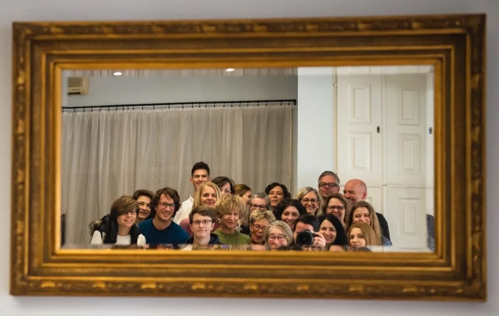 A group of people taking a photo together, reflected in a large rectangular mirror with a gold frame, in a room with white walls and curtains.