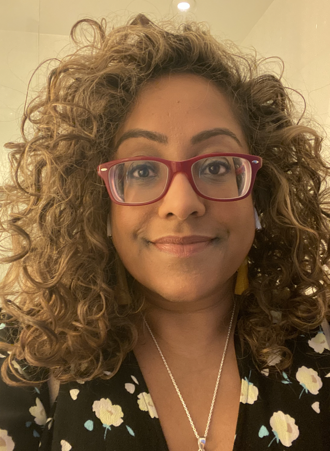 A woman with curly hair wearing red glasses, a floral top, and a silver necklace, smiling indoors.
