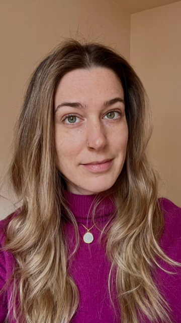 A woman with long wavy blonde hair wearing a magenta sweater and a gold necklace with a small round pendant, standing in front of a plain beige wall.