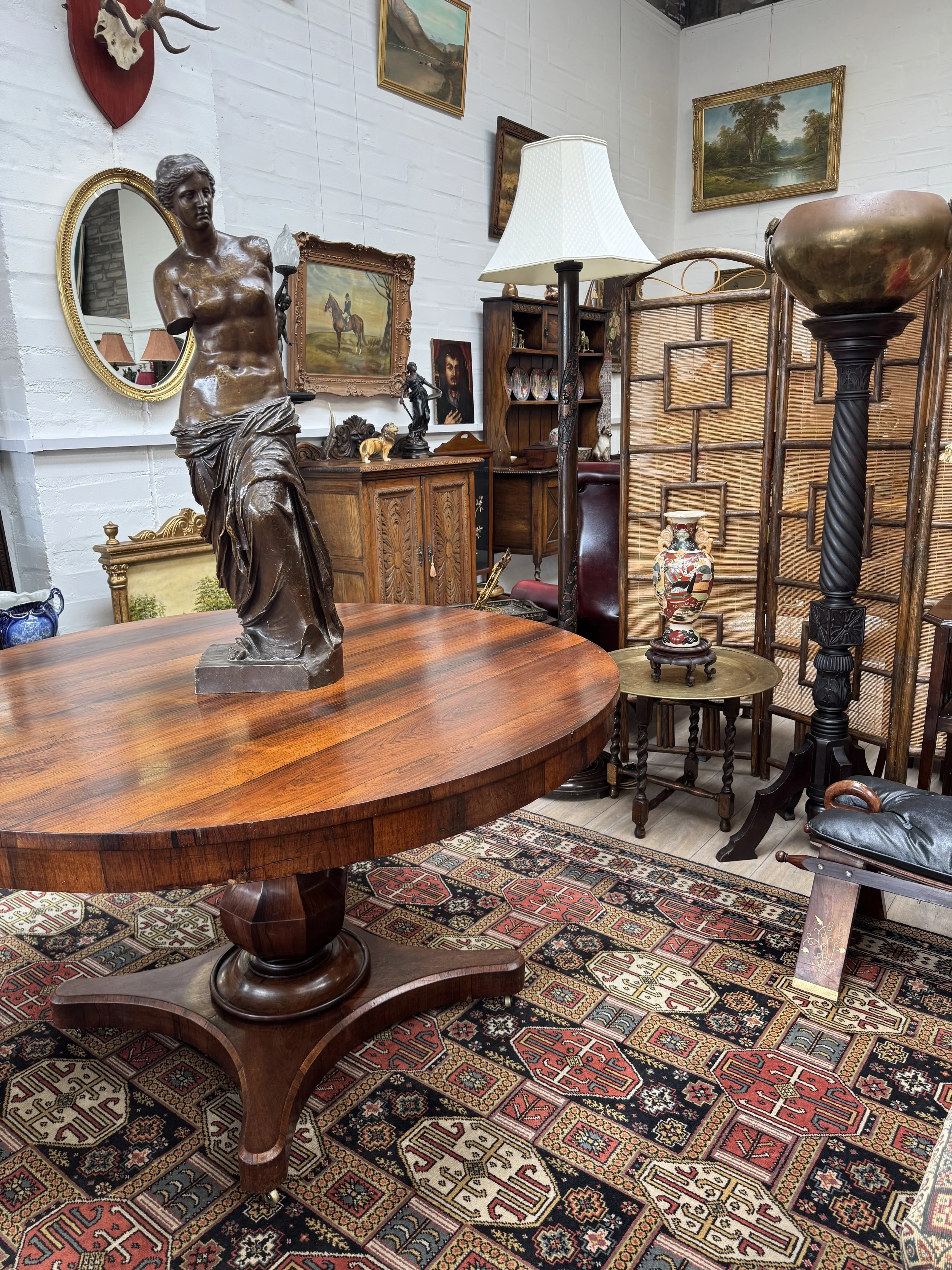 A vintage-style room decorated with various antique furniture and art. The centerpiece is a large, round wooden table with a bronze sculpture of a partially draped female figure on it. The room also features a Persian rug, a tall floor lamp, a wooden partition, framed landscape paintings, a small decorated vase, a mirror, and various small sculptures.