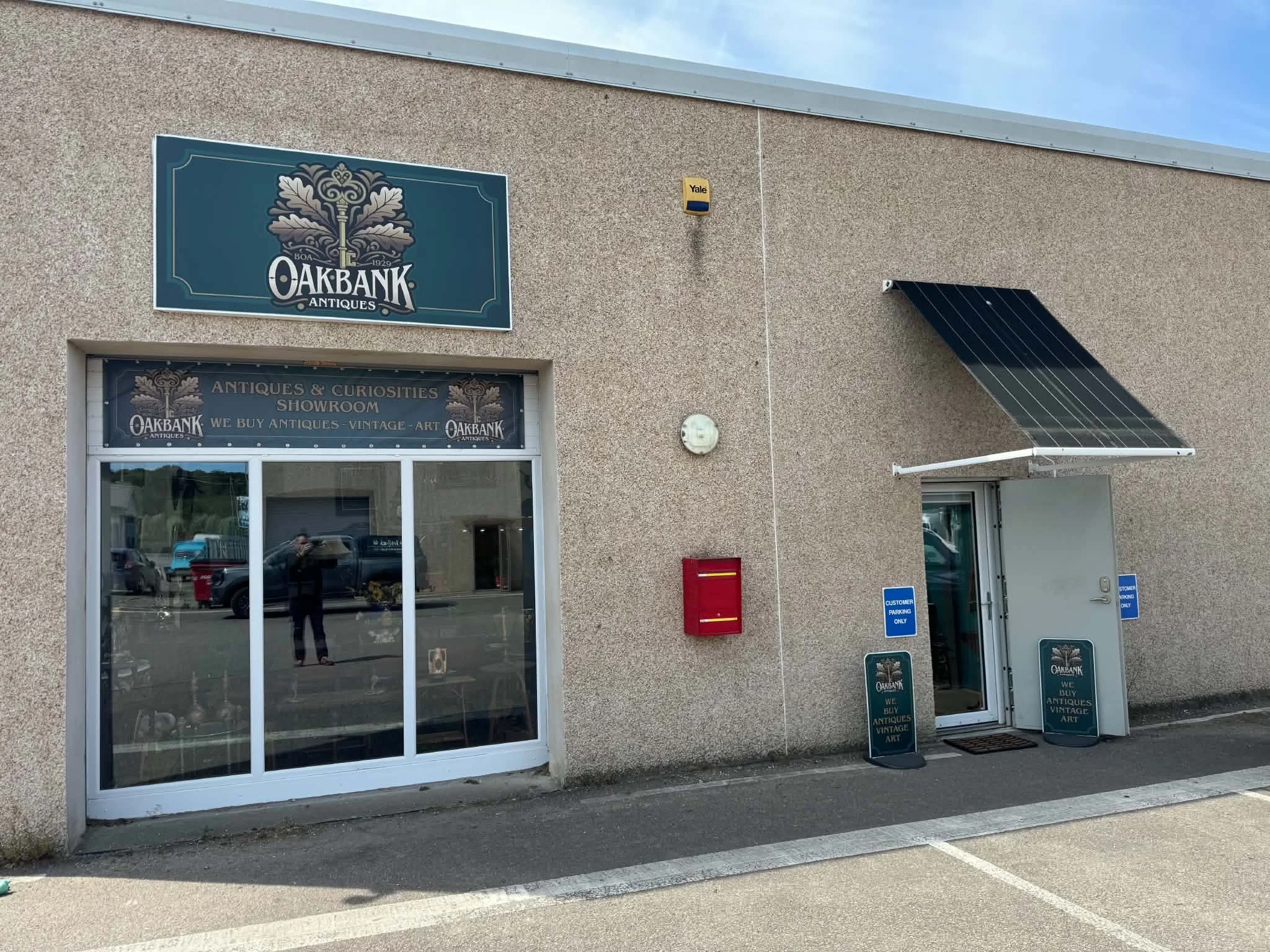 The exterior of a store named Oakbank Antiques, with large glass windows and signage indicating they buy antiques, vintage items, and art. There are two signs outside, one on the door and one on a stand, and a parking lot in front.