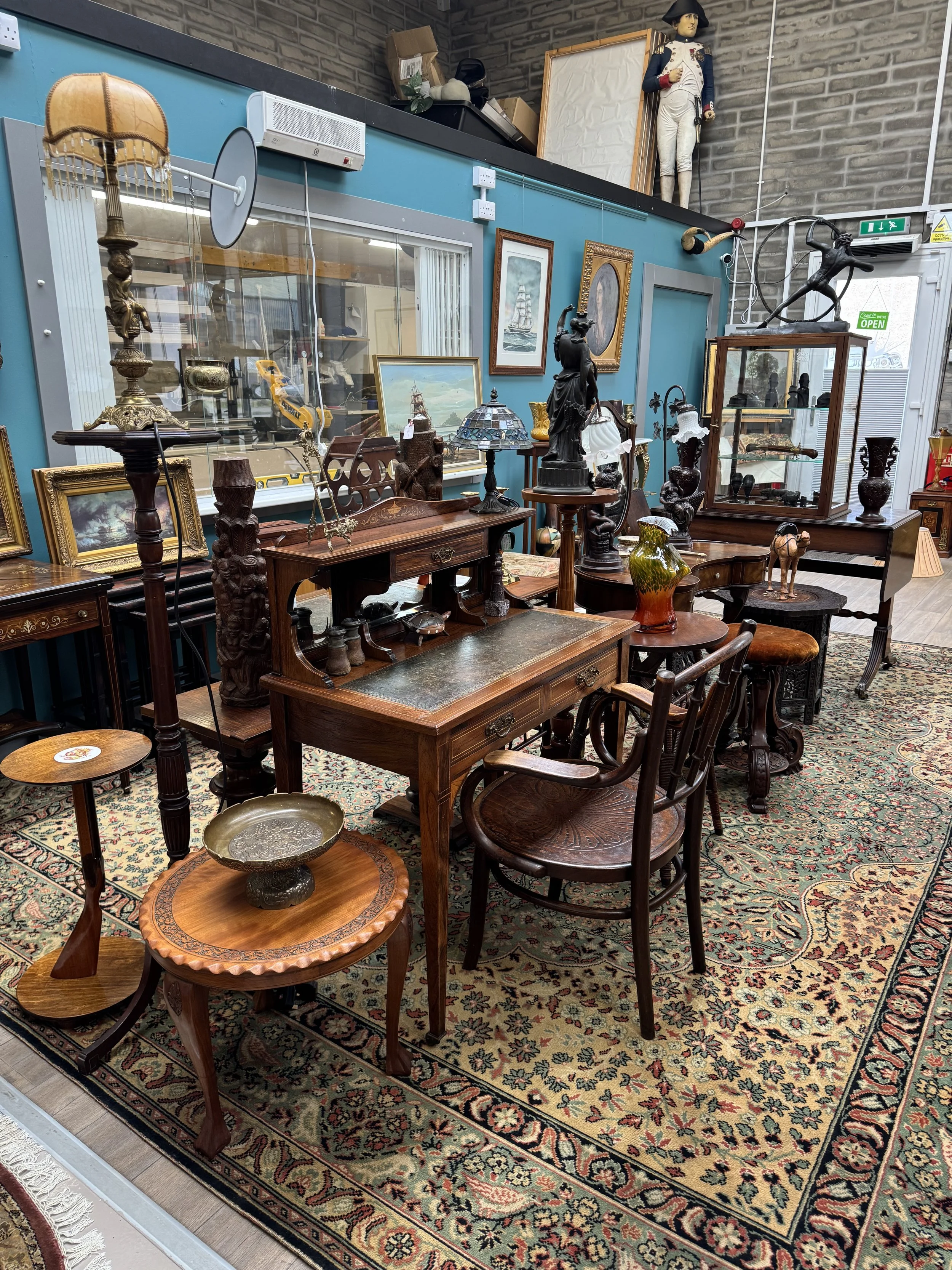 A collection of antique and vintage furniture and decorative items in a room, including wooden tables, chairs, lamps, paintings, sculptures, and a patterned area rug.