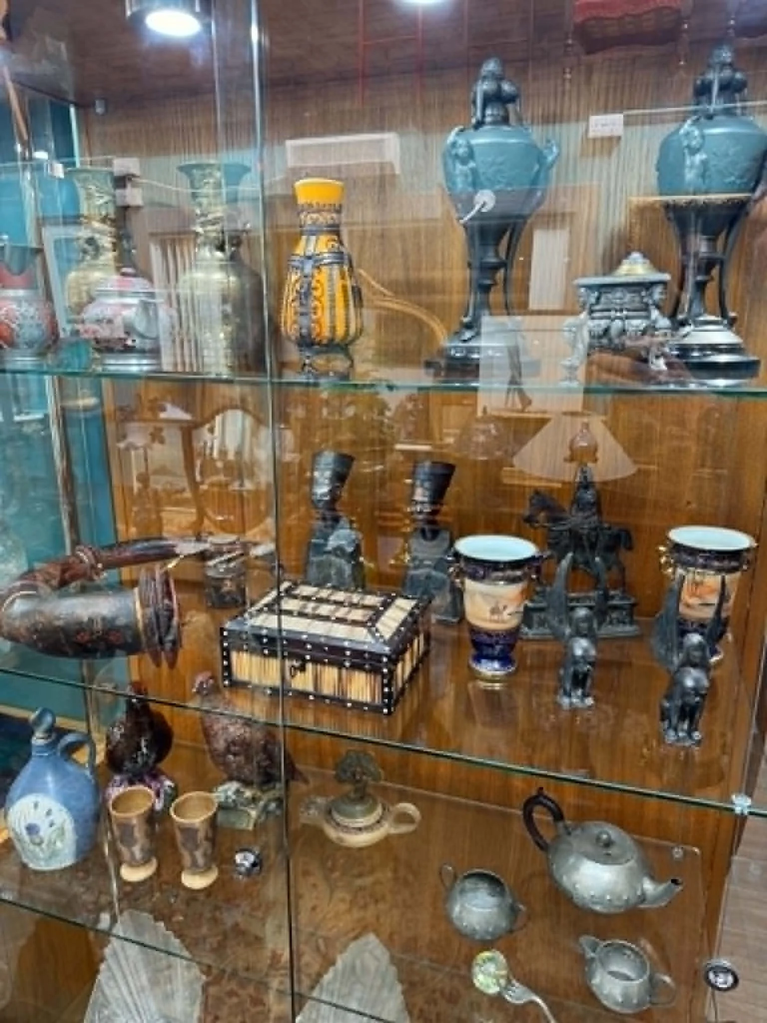 Display of antique ceramic and metalware items including vases, pots, lamps, and decorative objects arranged on glass shelves in a wooden display case.