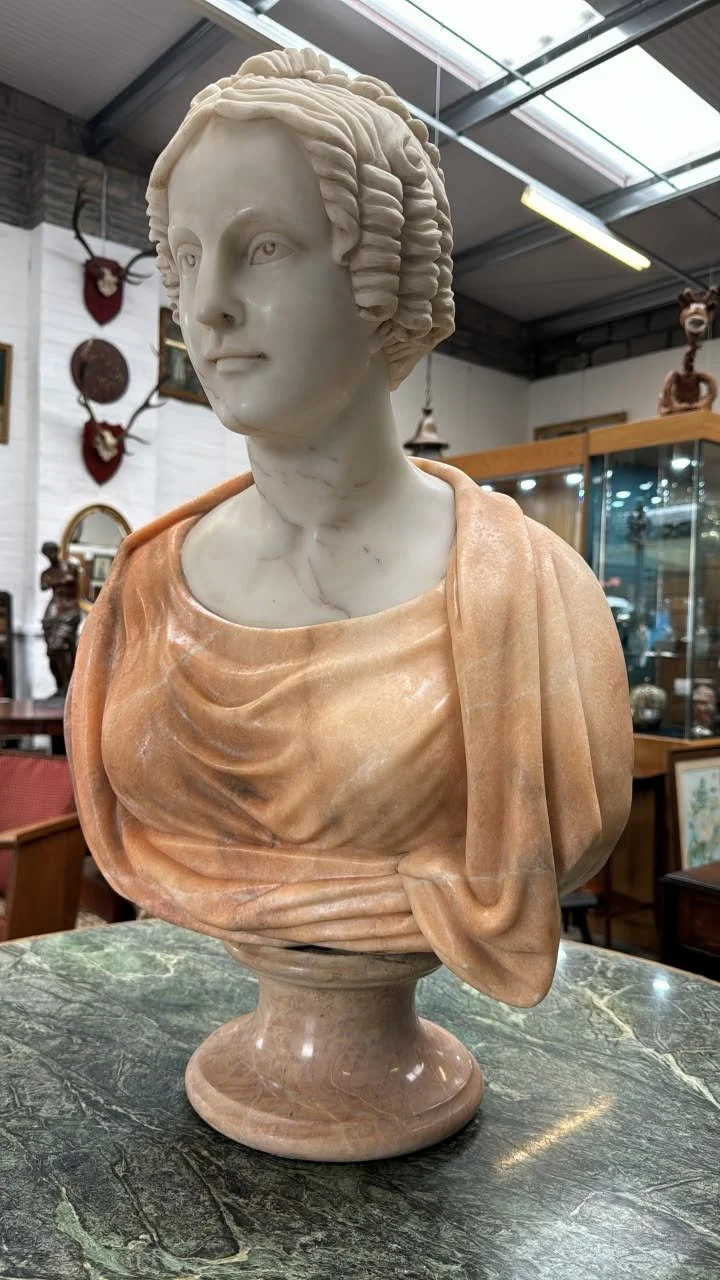 A marble bust of a woman with styled hair, dressed in draped clothing, displayed on a marble table inside a store with various decorative items in the background.