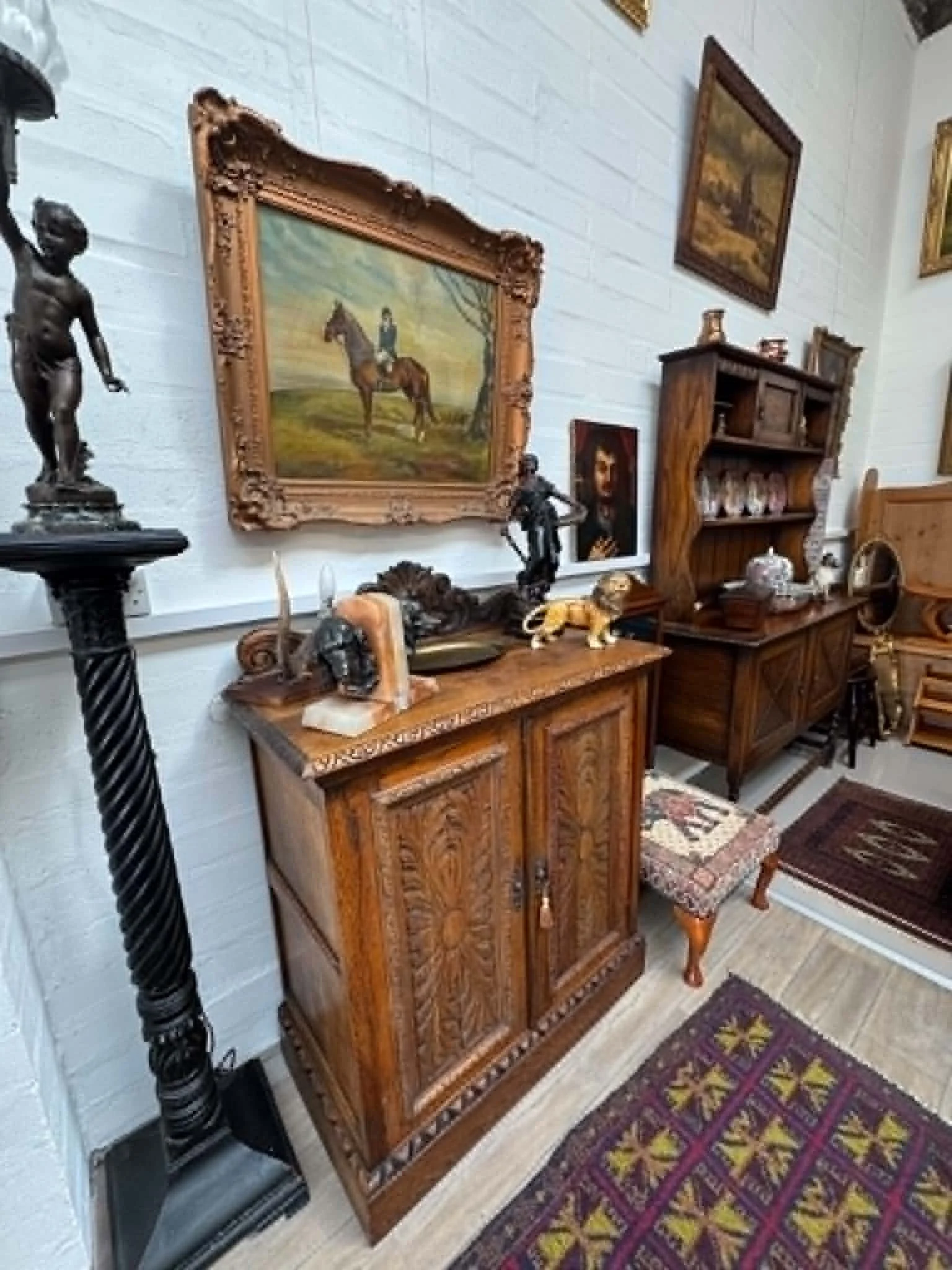 An interior room with antique wooden furniture, including a cabinet with carvings, a small table, and a decorative chair. There are various decorative items and artwork, including a large framed painting of a man on a horse, on the wall. The room also features a patterned rug on a wooden floor.