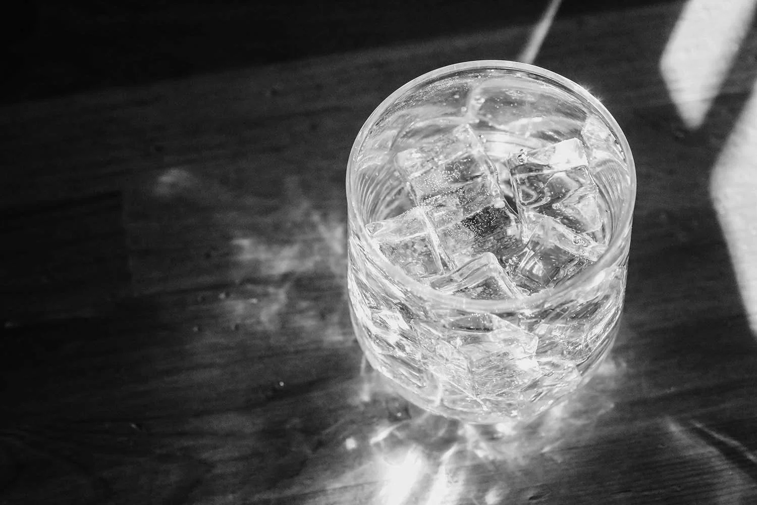 A glass filled with ice cubes and water, sitting on a dark wooden surface with shadows.