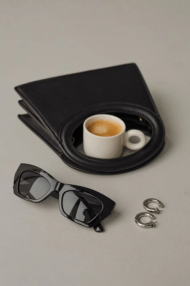A small white coffee cup with espresso inside, placed on a black leather wallet and lens case. Also includes a pair of black sunglasses and small silver hoop earrings.